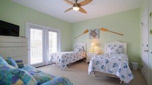 A bedroom with two beds, a ceiling fan, a TV, and a window with blinds. - Casa de la Playa: Spacious 4BR home in Holden Beach, perfect for family getaways. (Holden Beach)