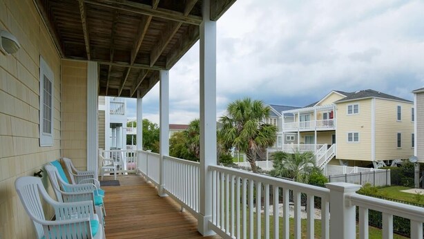 A covered porch with white railings and wooden flooring, leading to a residential area with houses and a palm tree. - Casa de la Playa: Spacious 4BR home in Holden Beach, perfect for family getaways. (Holden Beach)