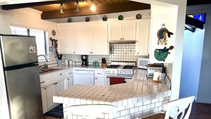 A kitchen with white cabinets, a stainless steel refrigerator, a gas stove, and a microwave. There are two white bar stools with woven seats.