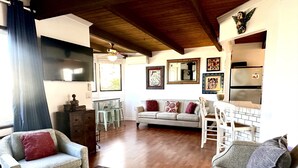 A spacious living room with a wooden ceiling, a sofa set, a dining area with a white table and chairs, and a kitchen with a refrigerator and a microwave.