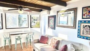 A cozy living room with a wooden ceiling, a gray sofa with decorative pillows, a dining area with a white table and green chairs, and artwork on the walls.
