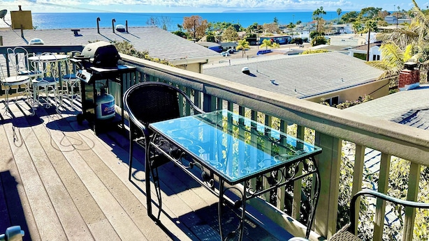 A balcony with a glass table and chairs overlooking a residential area and the ocean.