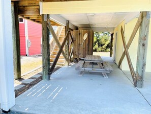 A covered outdoor area with a wooden table and benches, wooden beams, and a red building in the background. - Angling Inn / 111 Frigate: Spacious 4BR home in Holden Beach, perfect for adventures! (Holden Beach)