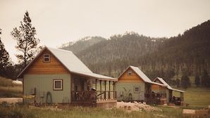 Two wooden cabins with porches, surrounded by a grassy field and a forested hillside.