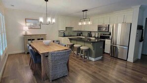 A modern kitchen with a central island, stainless steel appliances, and a dining area with a wooden table and wicker chairs. - Experience coastal bliss at Holden Haven, a spacious 4-bed retreat in Holden Beach. (Holden Beach)