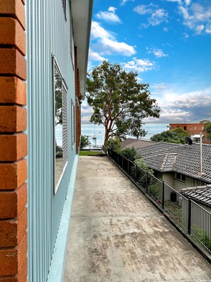 A balcony with a view of the sea, a tree, and a building with a tiled roof.