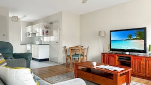 Ein Wohnzimmer mit Fernseher, der eine Strandansicht zeigt, einem Essbereich mit Stühlen, einer Couch und einem Couchtisch.