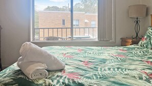 Uma cama com colcha florida, uma toalha branca enrolada e vista para um edifício através de uma janela.