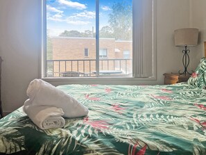 Een bed met een bloemenbedekker, een opgerolde witte handdoek en uitzicht op een gebouw door een raam.