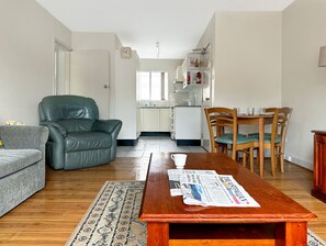 A living room with a sofa, a leather chair, a wooden dining table with chairs, and a kitchen in the background.