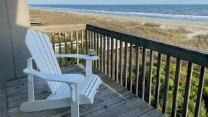 Ett trädäck med en vit Adirondack-stol och ett litet bord, med utsikt över en strand med sanddyner och havet.