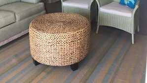 Una sala de estar con un sofá, dos sillones de mimbre, un otoman redondito y un ventilador de techo, con una cocina al fondo.