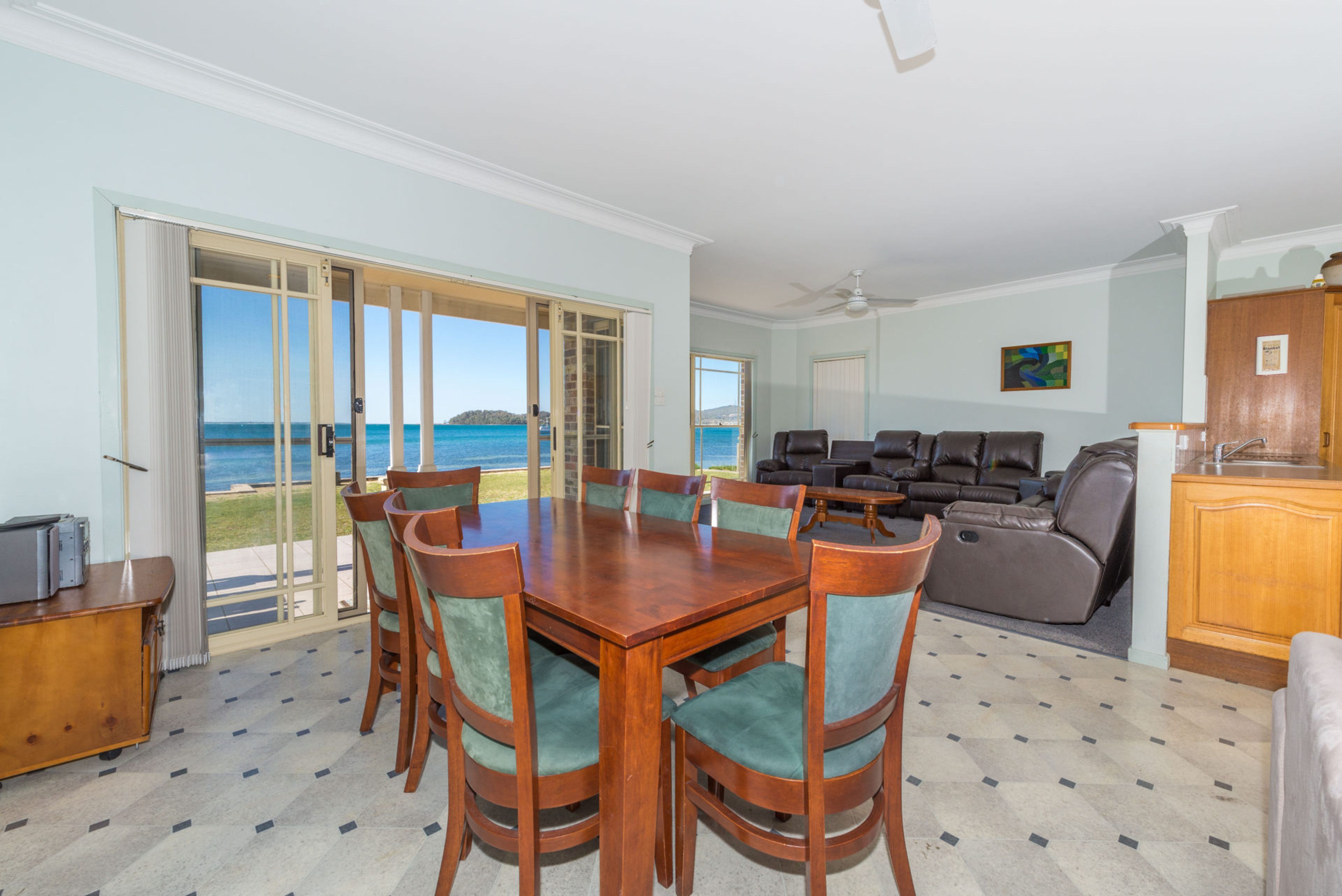 A spacious living area with a dining table, chairs, and a sofa. There is a view of the ocean through large glass doors.