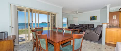 A spacious living area with a dining table, chairs, and a sofa. There is a view of the ocean through large glass doors.