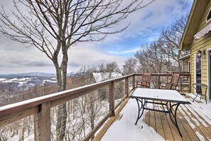 Una terraza de madera con una mesa y sillas, con vistas a un paisaje nevado con árboles desnudos y montañas distantes.