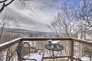Un balcón con una silla de metal y dos candelabros sobre una pequeña mesa, con vistas a un paisaje nevado con árboles desnudos y colinas a lo lejos.