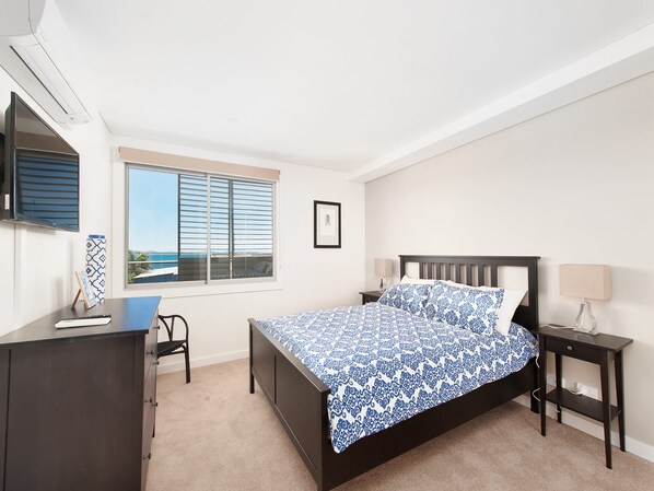 A bedroom with a bed, a desk, a chair, a window with blinds, and a wall-mounted TV.