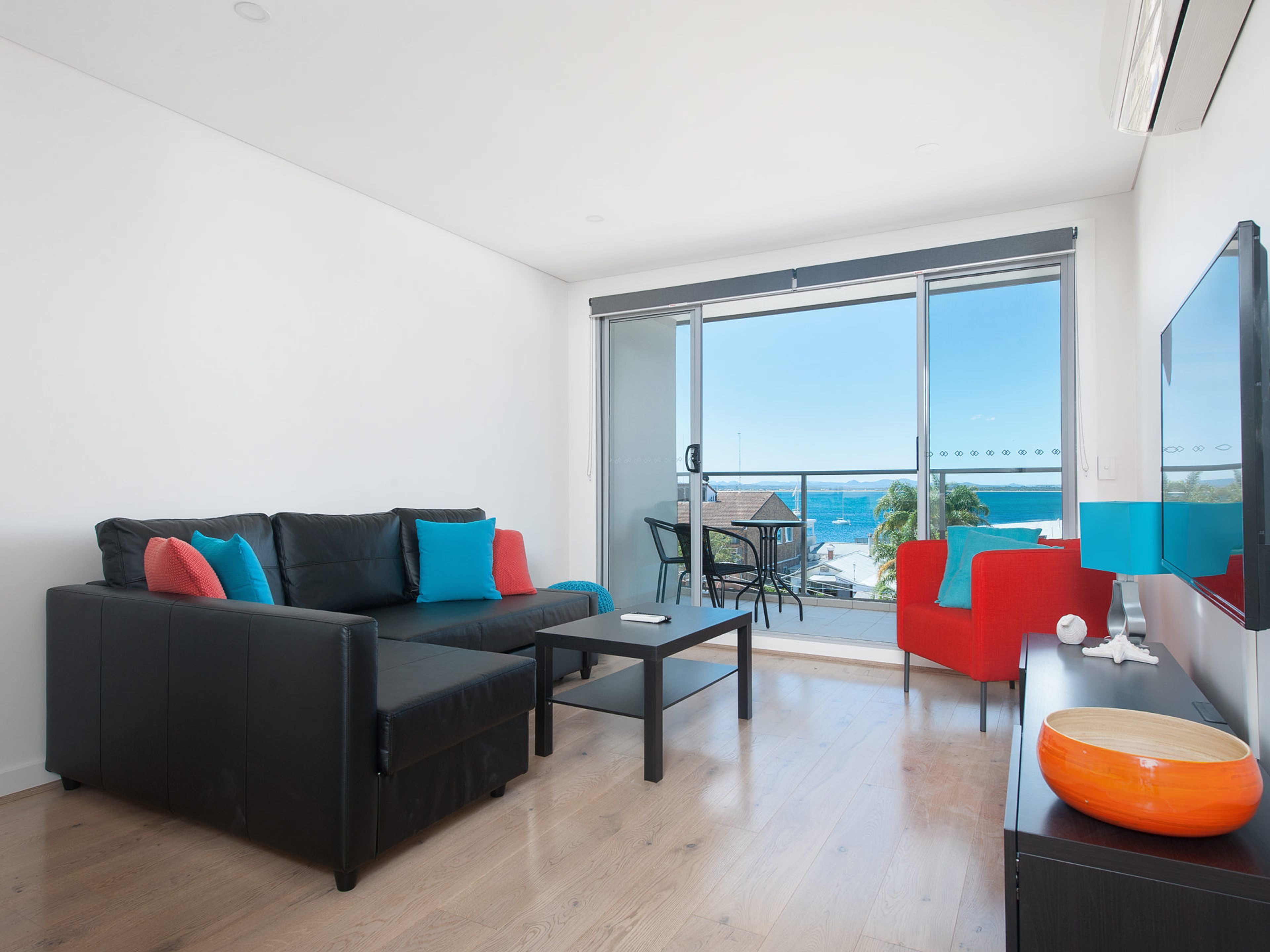 A modern living room with a black sofa, a small table, and a red armchair. A large window offers a view of the sea.