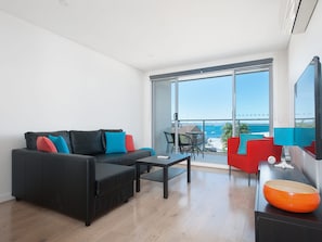Een moderne woonkamer met een zwarte bank, een klein tafeltje en een rode fauteuil. Een groot raam biedt uitzicht op zee.