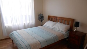 Un dormitorio con una cama de madera, una mesita de noche con lámpara, un ventilador de pie y una ventana con cortinas traslúcidas.