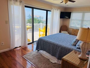A bedroom with a bed, a television, a ceiling fan, and a view of a balcony with yellow chairs.