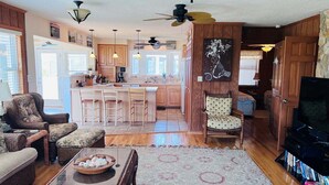 A spacious living room with a kitchen in the background, wooden paneling, a dining area with bar stools, and a television on a stand.