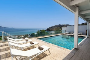 A swimming pool with lounge chairs, a clear view of the sea, and a hilly landscape in the background.