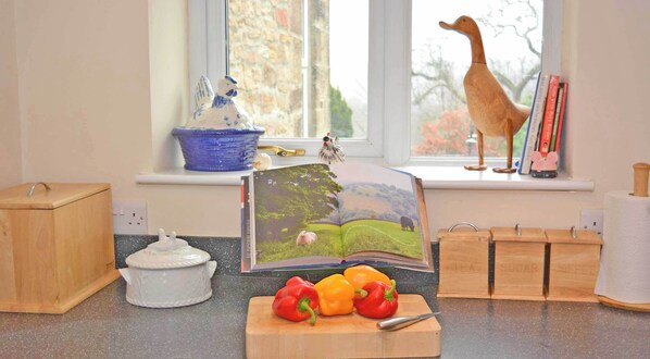 A kitchen counter with a book, a cutting board with peppers, a white ceramic pot, and wooden storage boxes.