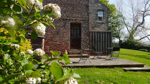 Un bâtiment rustique en pierre, doté d’une porte en bois, d’une petite fenêtre et d’un patio aménagé avec du mobilier d’extérieur.
