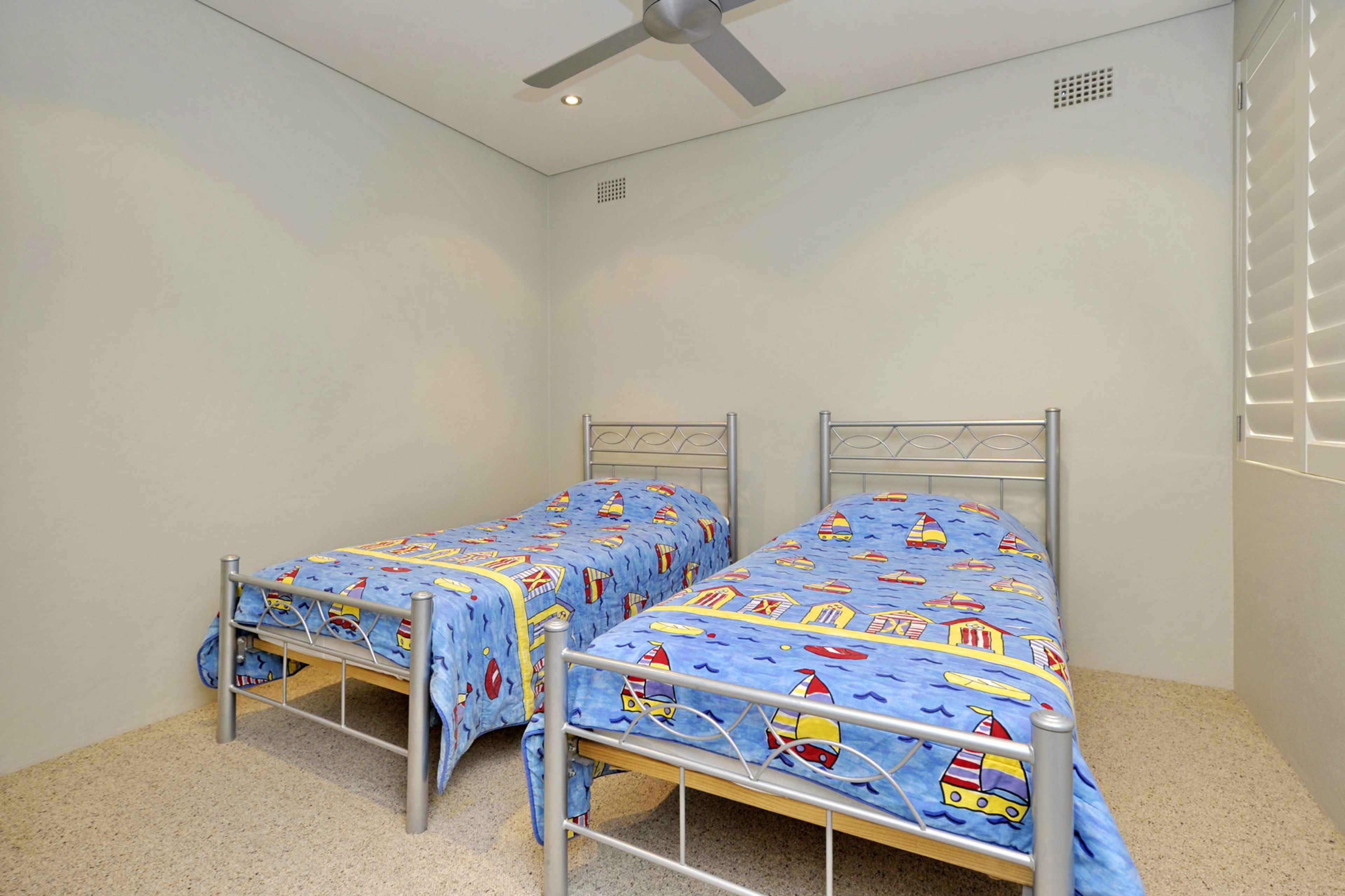 Un alojamiento pequeño y de una sola habitación, con dos camas de estructura metálica, un ventilador de techo y una ventana con persianas.