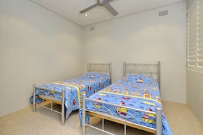 A small, single-room accommodation with two metal-framed beds, a ceiling fan, and a window with blinds.