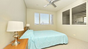 A bedroom with a bed, two bedside lamps, a ceiling fan, a window with blinds, and a carpeted floor. - Del Rae, Unit 12/25 Shoal Bay Road - North-facing apartment overlooking the Bay (Shoal Bay)