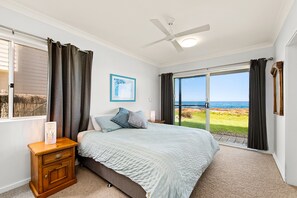 Een slaapkamer met een bed, nachtkastje, plafondventilator, raam met gordijnen en uitzicht op zee.