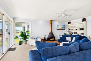 A modern living room with a fireplace, blue sofas, and a view through large glass doors. - Birubi Beach getaway at Anna Bay (Anna Bay)