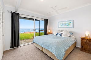 A bedroom with a large bed, a ceiling fan, a view of the ocean, and a wooden nightstand with a lamp. - Birubi Beach getaway at Anna Bay (Anna Bay)