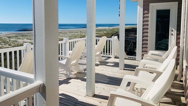 Een overdekt terras met witte stoelen, een houten terras en uitzicht op strand en zee.
