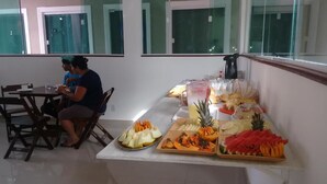 Una mesa de buffet con diversas frutas y una máquina de café. Se ven dos personas sentadas en una mesa al fondo.