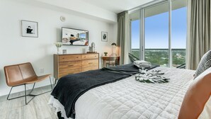 Quarto de hotel moderno com uma cama grande, cômoda de madeira, escrivaninha e uma cadeira. Há quadros nas paredes e vista da cidade através de janelas amplas.