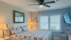 A bedroom with a bed, a ceiling fan, a window with blinds, a nightstand with a lamp, and a chair. - Enjoy oceanfront bliss at When Pigs Fly, a spacious 4BR gem in Holden Beach, NC. (Holden Beach)