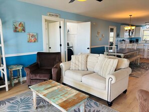 A living room with a brown armchair, a beige sofa, and a rustic coffee table. There is a door labeled 'seaside' and a kitchen area visible in the background.