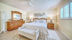 Une chambre à coucher comprenant un lit, des tables de chevet, une commode, un banc, un ventilateur de plafond et une fenêtre avec des stores.