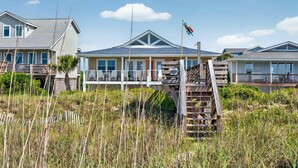 Ein Strandhaus mit überdachter Veranda, einer Holztreppe, die zu einer Terrasse führt, und einer Flagge auf einem Fahnenmast.