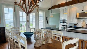Uma área de refeições com uma mesa de madeira, cadeiras brancas, um candelabro e vista para o exterior.