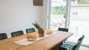 A wooden dining table with a white placemat, surrounded by green upholstered chairs. A fruit bowl with oranges and a pineapple is placed on the table. - Villa Lele - Pool, Sea & Mountain View (Novigrad)