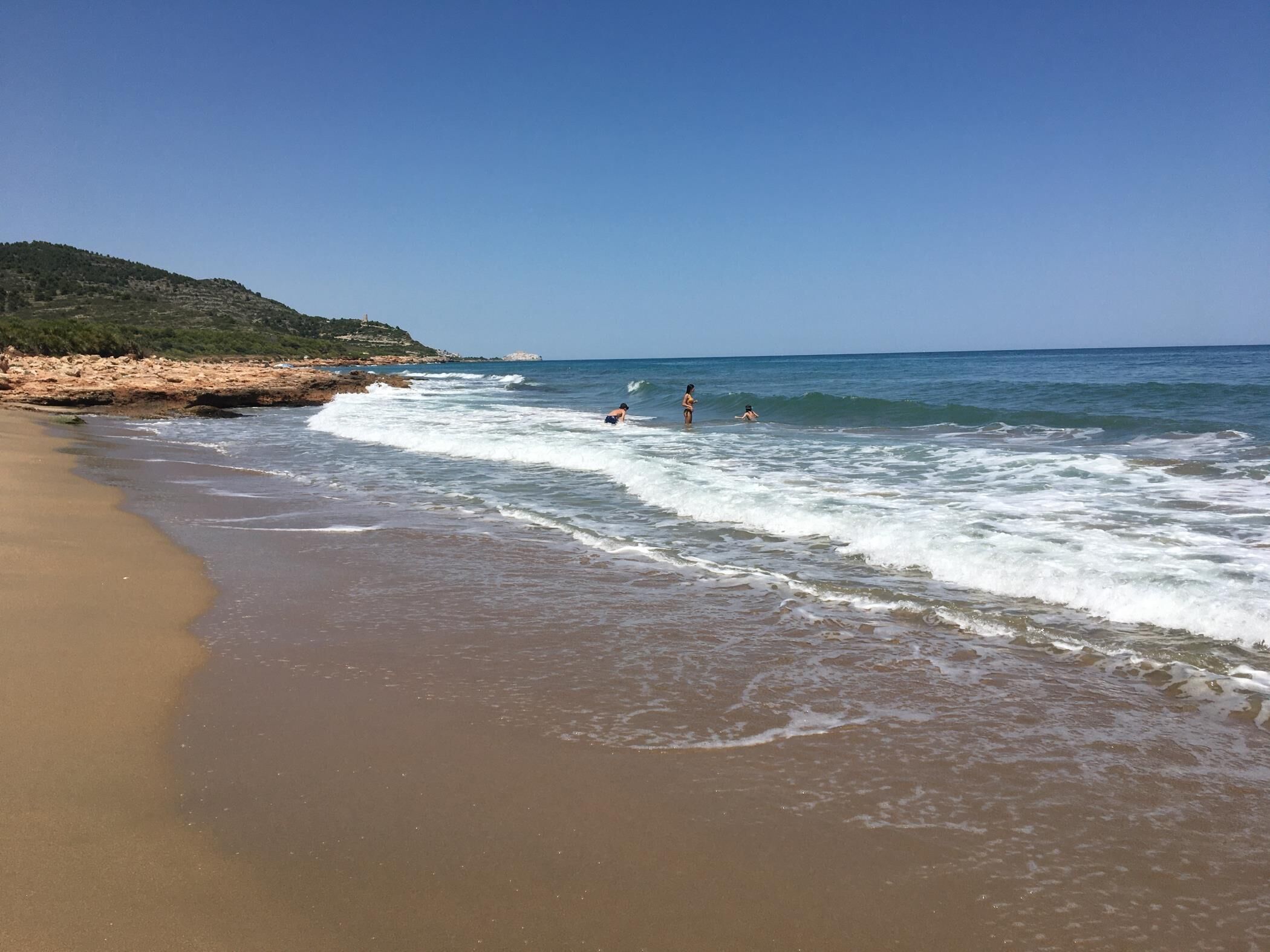 A beach with clear skies, waves, and a distant hill.