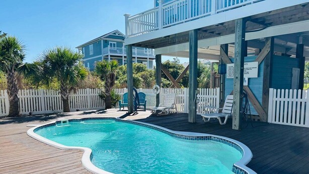 A swimming pool with a wooden deck, a white fence, and a building with a balcony. - Experience bliss at Blue Sunset in Holden Beach, NC! Sleeps 8. (Holden Beach)