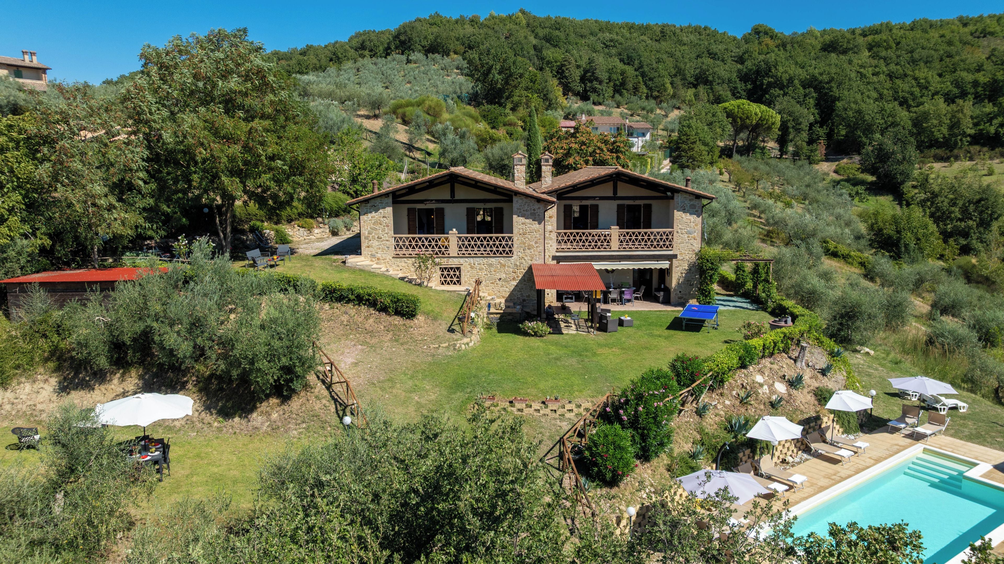 A two-story stone house with a pool, surrounded by a lush garden and hillside.
