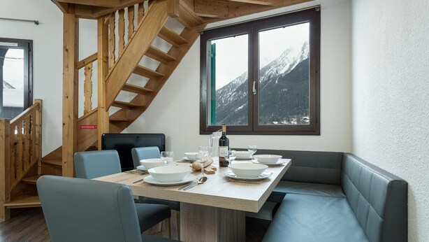 Een eethoek met een houten trap, een gedekte tafel met kommen en glazen, en een uitzicht op besneeuwde bergen door een raam.