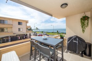 A balcony with a table and chairs, a grill, and a view of the sea and buildings.