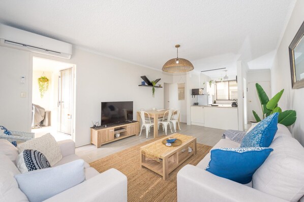 A modern living room with a TV, white sofas, a coffee table, and a kitchen in the background.